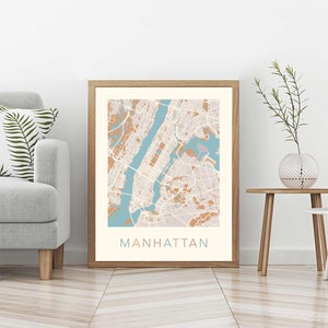 May include: A framed print of a map of Manhattan, New York City. The map is in a light brown frame and has a light blue and peach color scheme. The word "MANHATTAN" is printed in a light pink font at the bottom of the map.