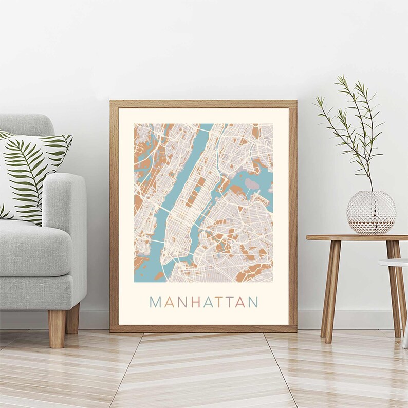 May include: A framed print of a map of Manhattan, New York City. The map is in a light brown frame and has a light blue and peach color scheme. The word "MANHATTAN" is printed in a light pink font at the bottom of the map.