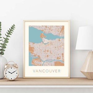 Op de afbeelding: Een ingelijste kaartprint van Vancouver, met een gedetailleerd stadsplan in beige en bruine tinten, met een lichtblauwe waterpartij. Het woord "VANCOUVER" is in een bijpassende kleur onderaan gedrukt.