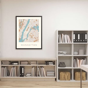 May include: A framed map of Manhattan, New York City, with a light brown and blue color scheme. The map is hanging on a white wall above a wooden bookshelf filled with books and decorative items.