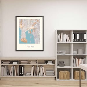 May include: A framed map of Tampa, Florida, with a light blue, brown, and gray color scheme. The map is hanging on a white wall above a wooden bookshelf filled with books and binders.