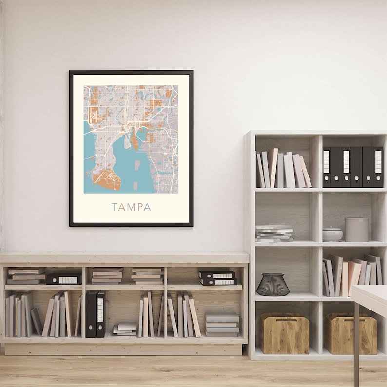 May include: A framed map of Tampa, Florida, with a light blue, brown, and gray color scheme. The map is hanging on a white wall above a wooden bookshelf filled with books and binders.