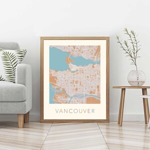 Op de afbeelding: Een ingelijste kaartprint van Vancouver, met een gedetailleerde stadsplattegrond in tinten beige, teal en bruin. Het woord "VANCOUVER" is in bijpassende teal kleur onderaan de print gedrukt. De print wordt weergegeven in een lichtbruine houten lijst.