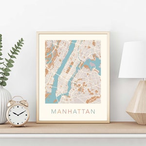 May include: A framed print of a map of Manhattan, New York City. The map is in a light blue, brown, and white color scheme. The word "MANHATTAN" is printed in a light pink, light blue, and brown color scheme at the bottom of the print.