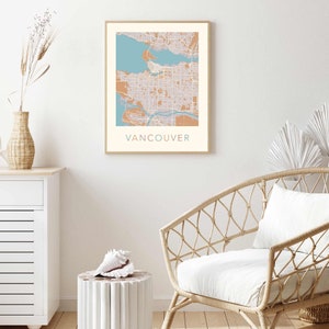 Op de afbeelding: Een ingelijste print met een kaart van Vancouver in tinten blauw, beige en bruin. De naam van de stad staat in turquoise onderaan afgedrukt. Het kunstwerk wordt tentoongesteld in een kamer met een witte muur, een rotan stoel en een decoratieve vaas.