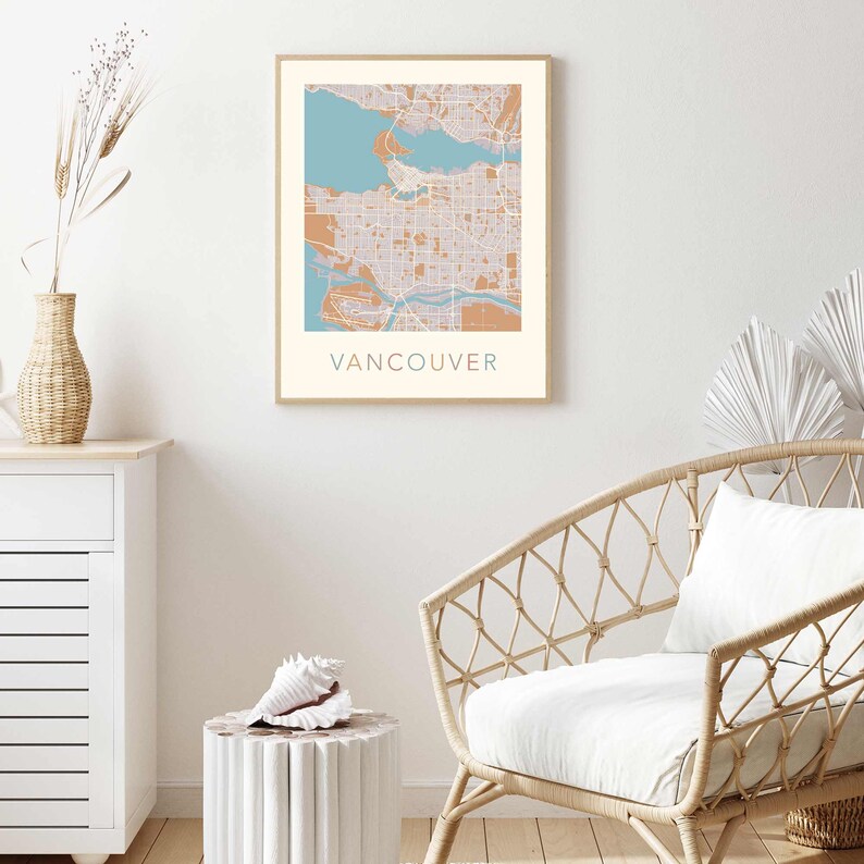 Op de afbeelding: Een ingelijste print met een kaart van Vancouver in tinten blauw, beige en bruin. De naam van de stad staat in turquoise onderaan afgedrukt. Het kunstwerk wordt tentoongesteld in een kamer met een witte muur, een rotan stoel en een decoratieve vaas.