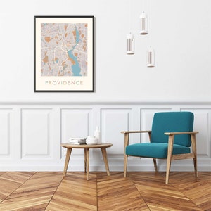 May include: A teal armchair with wooden legs sits in a room with white walls and wood floors. A framed map of Providence, Rhode Island hangs on the wall above a wooden coffee table with books and white ceramic vases.