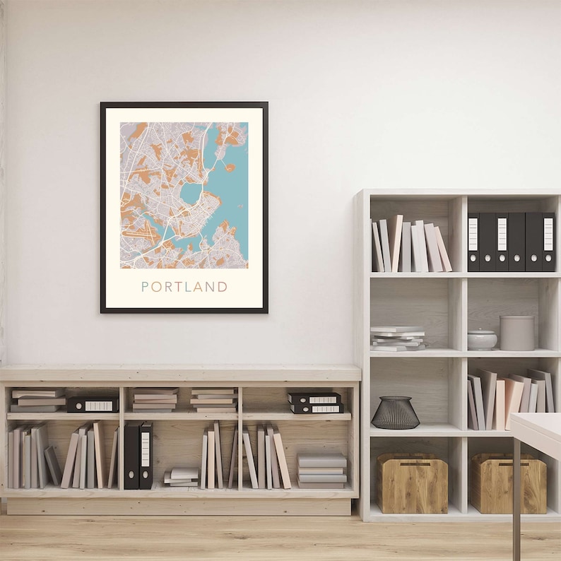 Pu&ograve; includere: Una mappa incorniciata di Portland, Maine, con uno schema di colori blu chiaro e arancione. La mappa &egrave; appesa a un muro bianco sopra una libreria in legno piena di libri e raccoglitori.