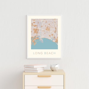 May include: A framed print of a map of Long Beach, California. The map is in shades of brown and blue, with the city streets and coastline outlined in white. The city name is printed in a light blue font at the bottom of the map.