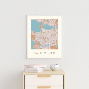 Op de afbeelding: Een ingelijste kunstprint van Vancouver, met een gedetailleerde stadskaart in gedempte tinten beige, blauw en bruin. Het woord "VANCOUVER" staat onderaan afgedrukt. De print wordt tentoongesteld aan een witte muur boven een houten kast.