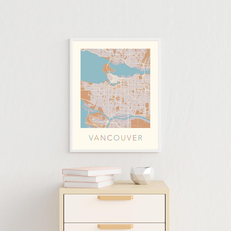Op de afbeelding: Een ingelijste kunstprint van Vancouver, met een gedetailleerde stadskaart in gedempte tinten beige, blauw en bruin. Het woord "VANCOUVER" staat onderaan afgedrukt. De print wordt tentoongesteld aan een witte muur boven een houten kast.