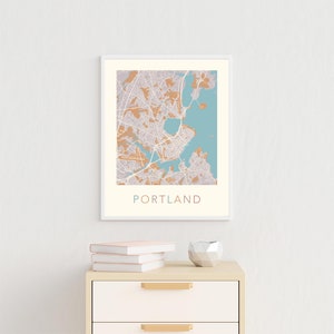 Pu&ograve; includere: Una stampa di mappa incorniciata di Portland, Maine, con uno schema di colori blu chiaro e beige. La mappa &egrave; esposta su una parete bianca sopra una cassettiera beige chiaro con maniglie dorate. Ci sono tre libri impilati sopra la cassettiera.