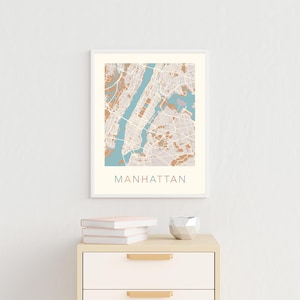 May include: A framed print of a map of Manhattan, New York City. The map is in shades of brown and blue, with the city streets and landmarks outlined in white. The word "MANHATTAN" is printed in a colorful font below the map.