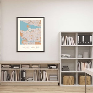Op de afbeelding: Een ingelijste kaartprint van Vancouver, met een gedetailleerde stadsplattegrond in tinten beige, blauw en oranje. Het woord "VANCOUVER" staat onderaan afgedrukt. De print wordt in een zwarte lijst aan een witte muur getoond.
