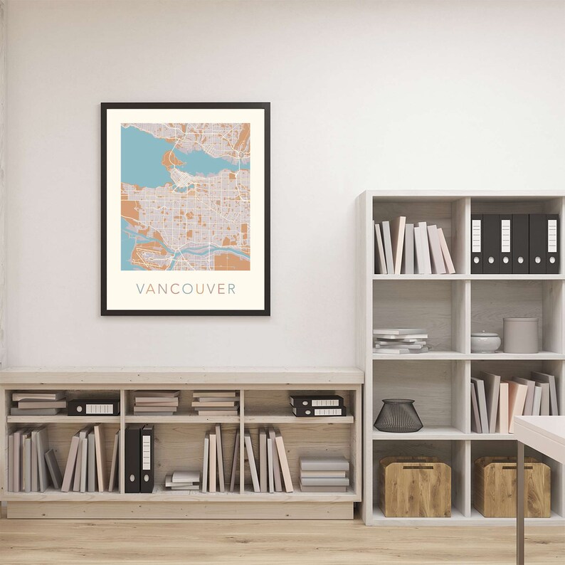Op de afbeelding: Een ingelijste kaartprint van Vancouver, met een gedetailleerde stadsplattegrond in tinten beige, blauw en oranje. Het woord "VANCOUVER" staat onderaan afgedrukt. De print wordt in een zwarte lijst aan een witte muur getoond.