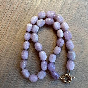 May include: A necklace featuring large, rounded, lavender-colored beads. The beads are strung together and secured with a gold-colored clasp. The necklace is displayed on a light-colored wooden surface.