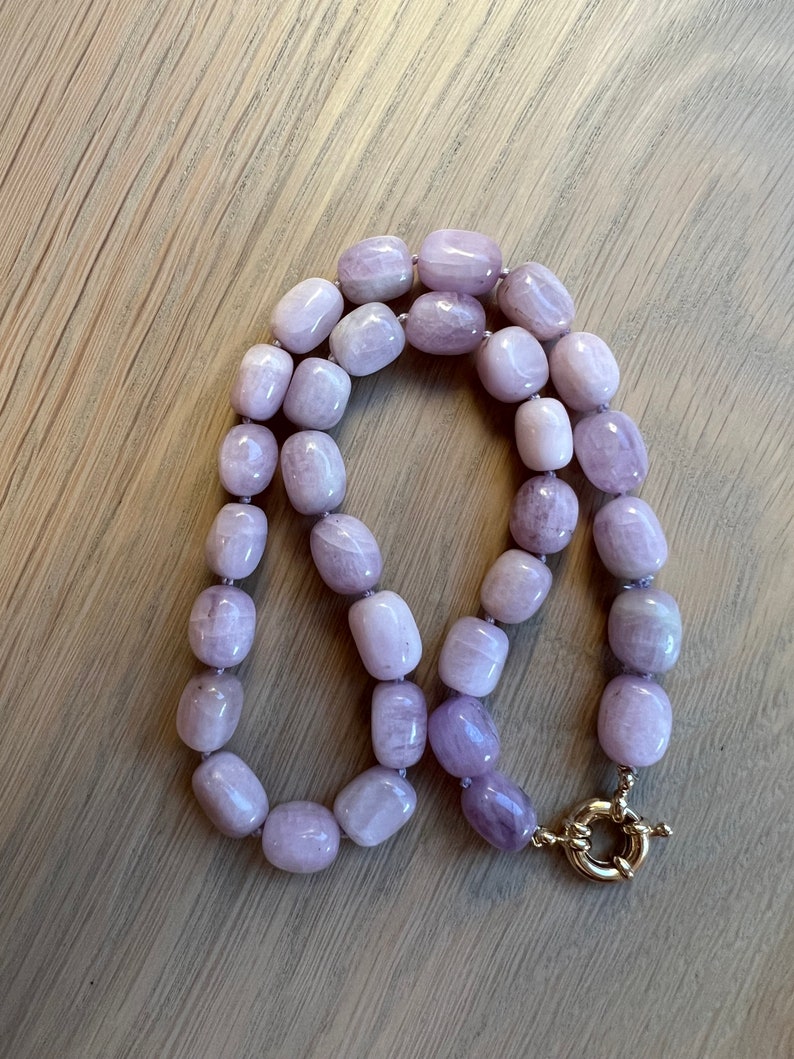 May include: A necklace featuring large, rounded, lavender-colored beads. The beads are strung together and secured with a gold-colored clasp. The necklace is displayed on a light-colored wooden surface.