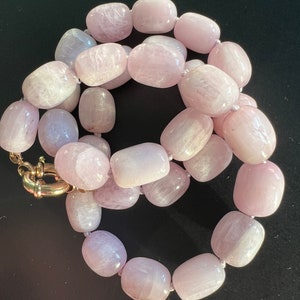 May include: A necklace featuring large, oval-shaped, light pink beads. The beads are strung together, creating a continuous loop. The clasp is gold-colored. The necklace is displayed against a dark background.