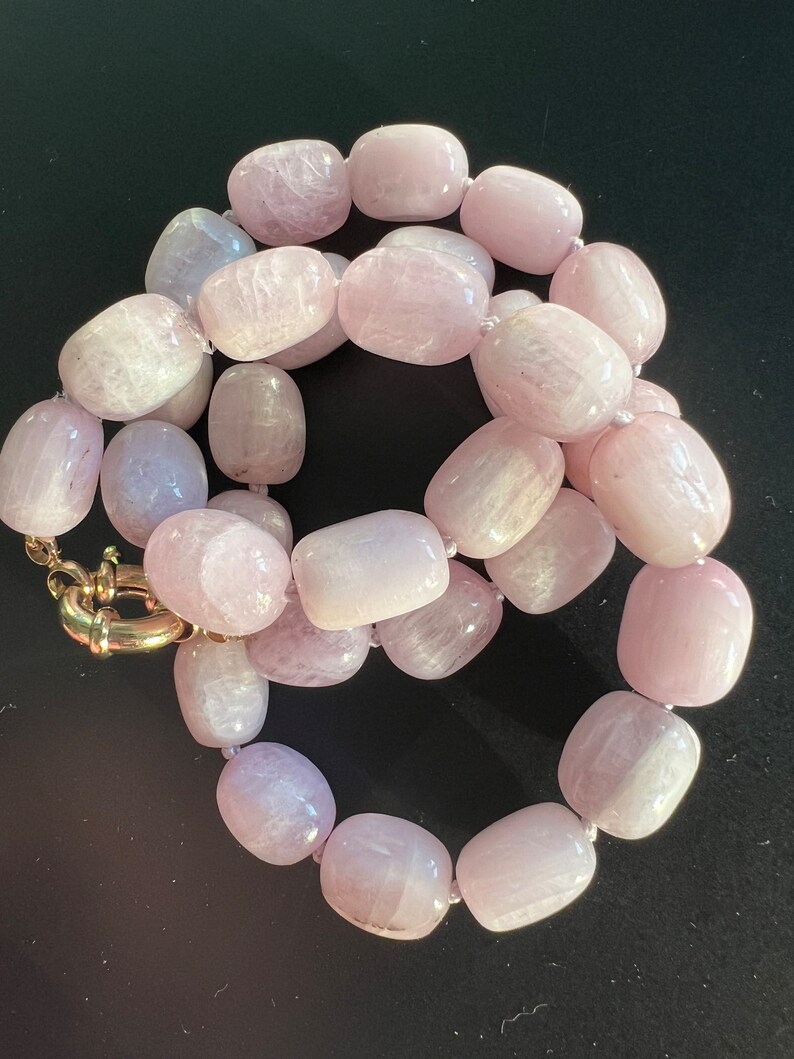 May include: A necklace featuring large, oval-shaped, light pink beads. The beads are strung together, creating a continuous loop. The clasp is gold-colored. The necklace is displayed against a dark background.