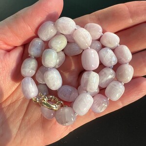 May include: A necklace featuring light purple, oval-shaped beads. The beads are strung together with a gold-colored clasp. The necklace is displayed against a dark background.
