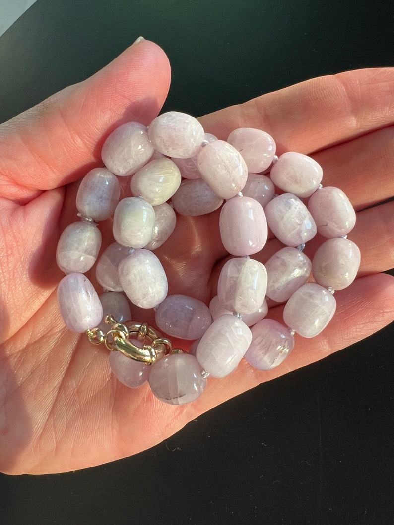 May include: A necklace featuring light purple, oval-shaped beads. The beads are strung together with a gold-colored clasp. The necklace is displayed against a dark background.