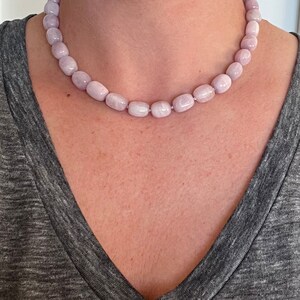 May include: A light purple beaded necklace. The necklace features smooth, oval-shaped beads strung together. The beads have a soft, pastel color and a subtle sheen. The necklace is worn around the neck, resting on the chest.