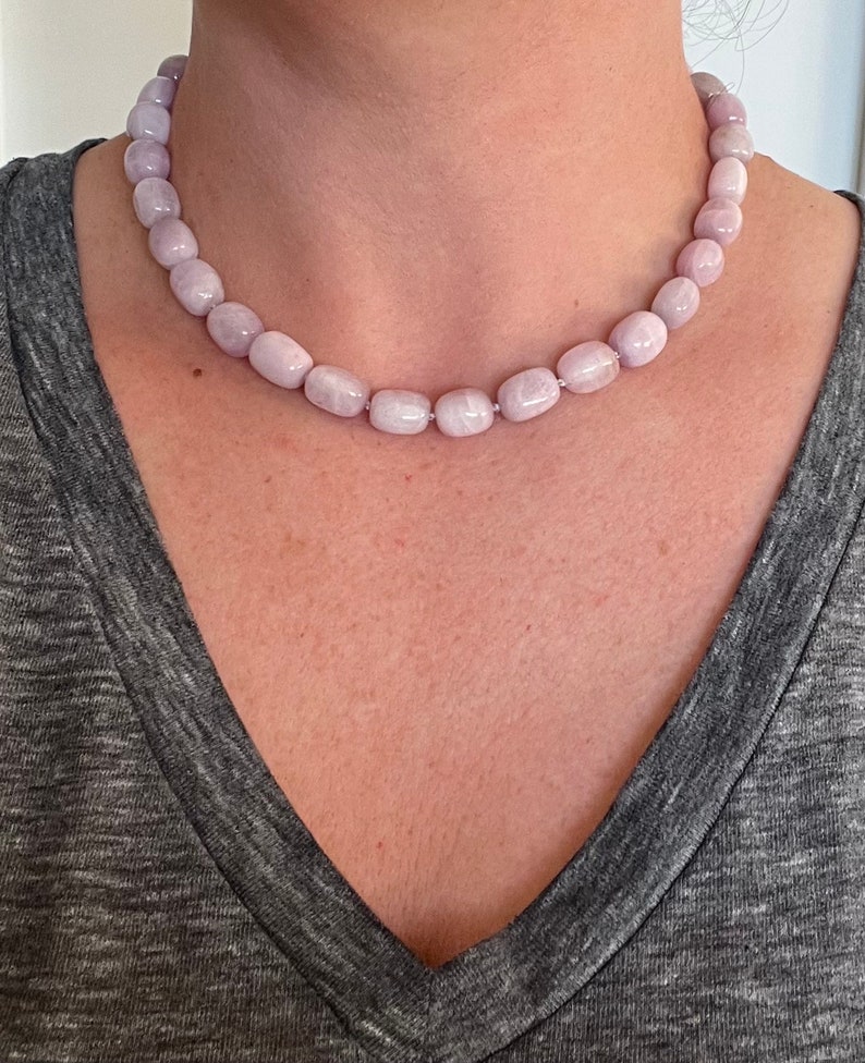 May include: A light purple beaded necklace. The necklace features smooth, oval-shaped beads strung together. The beads have a soft, pastel color and a subtle sheen. The necklace is worn around the neck, resting on the chest.
