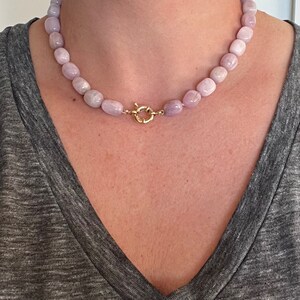 May include: A light purple beaded necklace with a gold-colored clasp. The necklace is composed of rounded, light purple beads. The clasp is a circular design with a secure closure. The necklace is worn against a gray shirt.