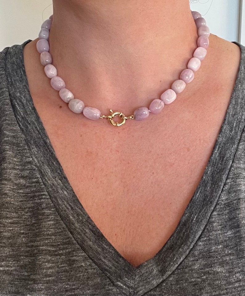 May include: A light purple beaded necklace with a gold-colored clasp. The necklace is composed of rounded, light purple beads. The clasp is a circular design with a secure closure. The necklace is worn against a gray shirt.