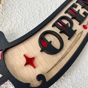 Könnte beinhalten: Ein dekoratives Holzschild mit einer hellen Holzbasis, einem schwarzen Rand und dem schwarzen Schriftzug OPEN. Das Design enthält eingelegte, sternförmige Akzente aus rotem Acryl und eine goldene Kette an der Oberseite zum Aufhängen.