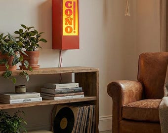 May include: A red rectangular wall lamp with the word "CONO!" in orange letters. The lamp is mounted on a light-coloured wall, with a string of lights above. A wooden bookshelf with plants and books is in the foreground.