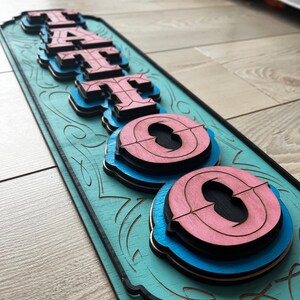 May include: A wooden sign with the word "TATTOO" in layered, 3D letters. The letters are painted in shades of pink, blue, and black, and the background is a light teal color. The sign has a decorative border with a floral design.
