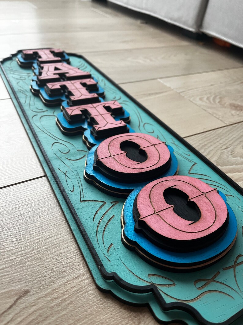 May include: A wooden sign with the word "TATTOO" in layered, 3D letters. The letters are painted in shades of pink, blue, and black, and the background is a light teal color. The sign has a decorative border with a floral design.