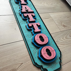 May include: A wooden sign with the word "TATTOO" in pink and blue letters. The sign is painted a light blue color and has a decorative border.