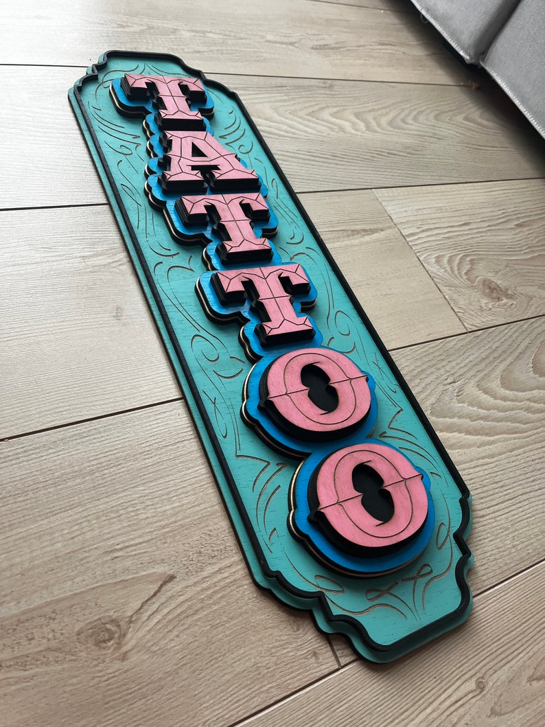 May include: A wooden sign with the word "TATTOO" in pink and blue letters. The sign is painted a light blue color and has a decorative border.