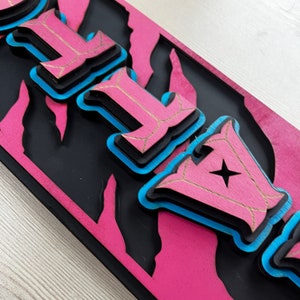 以下が含まれることがあります： ピンクと黒のウッドサインに、レイヤー状の3Dスタイルで「FLY」の文字が書かれています。文字は青で縁取られ、金色に仕上げられています。