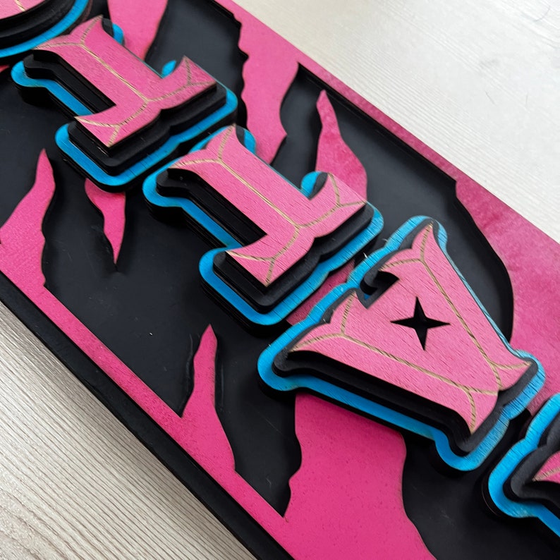 以下が含まれることがあります： ピンクと黒のウッドサインに、レイヤー状の3Dスタイルで「FLY」の文字が書かれています。文字は青で縁取られ、金色に仕上げられています。
