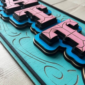 May include: A wooden sign with the word "HAUTE" in pink and blue layered letters. The sign has a blue background with a black border and a decorative swirl pattern.