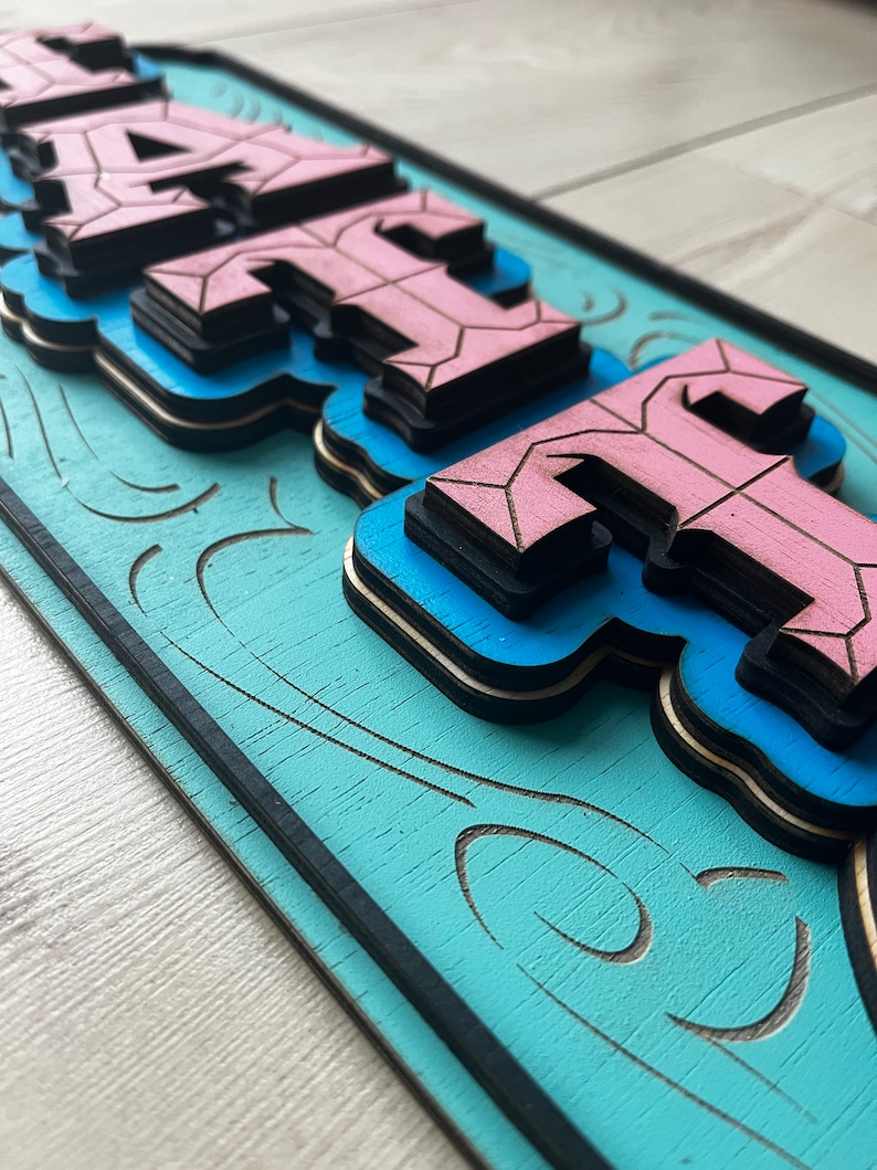 May include: A wooden sign with the word "HAUTE" in pink and blue layered letters. The sign has a blue background with a black border and a decorative swirl pattern.