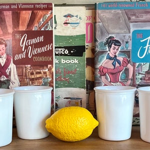 May include: Four white, cylindrical glasses are arranged in front of vintage cookbooks, including "The German and Viennese Cookbook" and "The French Cookbook." A bright yellow lemon sits between the glasses. The cookbooks feature illustrations of women in kitchens.