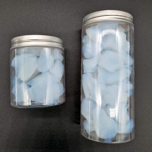 Peut inclure: Deux bocaux en plastique transparent avec des couvercles argentés, remplis de pétales bleu clair. Les bocaux sont de tailles différentes, le plus grand étant plus haut que le plus petit. Les pétales semblent être faits d'une matière douce.