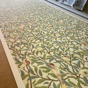 May include: A cream-coloured wallpaper with a repeating pattern of green leaves, brown branches, and yellow birds. The birds are perched on the branches and have red accents on their chests. The wallpaper has a vintage aesthetic.