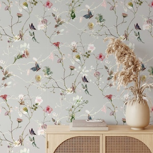 Puede incluir: Un papel tapiz gris con un patrón floral que presenta ramas delicadas, flores rosas y blancas y mariposas azules. El papel tapiz se muestra en una pared detrás de un armario de madera con un frente de mimbre.