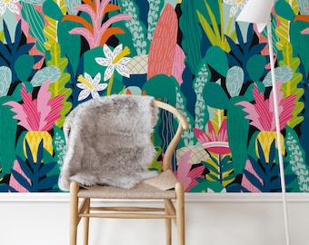 Op de afbeelding: Een kleurrijk tropisch behang met een verscheidenheid aan groene, roze, gele en blauwe bladeren en bloemen. Een houten stoel met een grijs kunstbont kussen staat voor het behang. Een witte lamp met een witte kap staat rechts in de afbeelding.