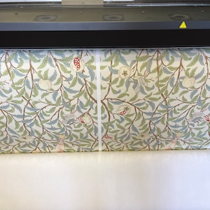 May include: A large format printer is printing a roll of fabric with a repeating pattern of stylised birds, pomegranates, and leaves in shades of green, blue, red, and brown on a white background.