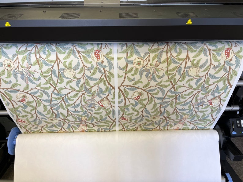 May include: A large format printer is printing a roll of fabric with a repeating pattern of stylised birds, pomegranates, and leaves in shades of green, blue, red, and brown on a white background.