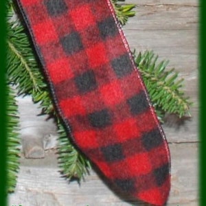 May include: A close-up of a red and black plaid ribbon, possibly for a wreath or holiday decoration. The ribbon is made of a soft material and is surrounded by green pine needles. The text "Maine Balsam Wreaths" is at the bottom.