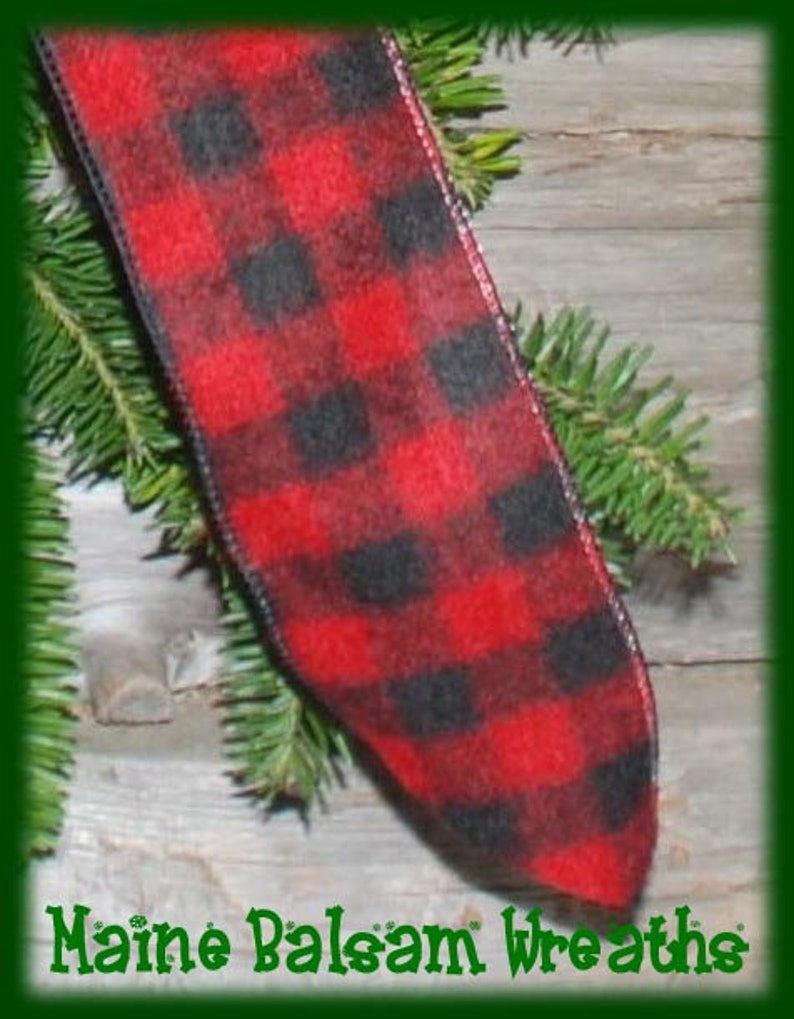 May include: A close-up of a red and black plaid ribbon, possibly for a wreath or holiday decoration. The ribbon is made of a soft material and is surrounded by green pine needles. The text "Maine Balsam Wreaths" is at the bottom.