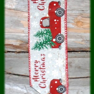 May include: A Christmas-themed ribbon with a red truck carrying a Christmas tree. The ribbon has the words "Merry Christmas" and snowflakes on a white background. The edges are red.