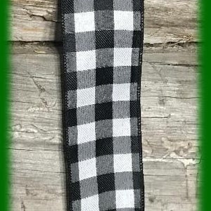 May include: A black and white checkered ribbon with a white button at the top. The ribbon is displayed vertically against a wooden background. The text "Maine Balsam Wreaths" is at the bottom.